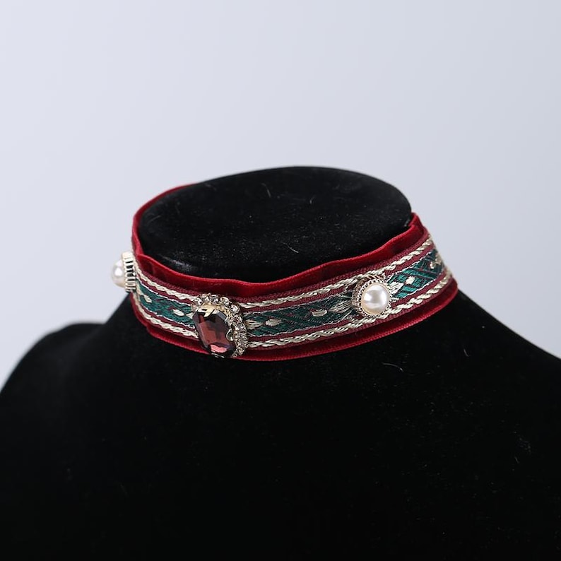 May include: A burgundy velvet choker necklace with a green and gold patterned ribbon. The choker features a large, oval, purple gemstone and two pearls. The necklace is displayed on a black mannequin head.