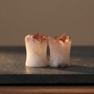May include: A pair of ear plugs crafted from translucent white stone with jagged, crystalline tops in shades of amber and brown. The cylindrical bodies feature a smooth, concave shape, showcasing natural mineral textures and light-catching edges.