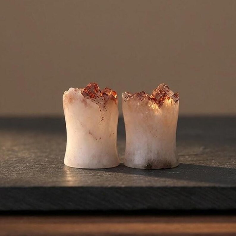 May include: A pair of ear plugs crafted from translucent white stone with jagged, crystalline tops in shades of amber and brown. The cylindrical bodies feature a smooth, concave shape, showcasing natural mineral textures and light-catching edges.