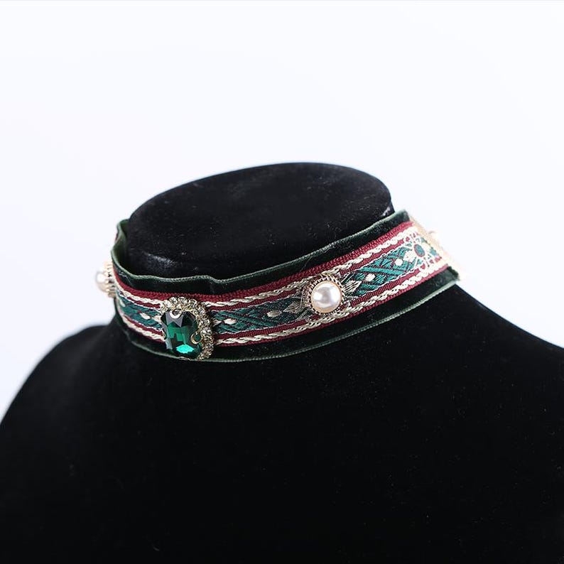 Pode incluir: Um colar gargantilha de veludo verde com uma faixa decorativa com um padr&atilde;o geom&eacute;trico em vermelho e verde. A gargantilha &eacute; adornada com uma grande gema de cor esmeralda e p&eacute;rolas falsas. O colar &eacute; exibido em um manequim preto.