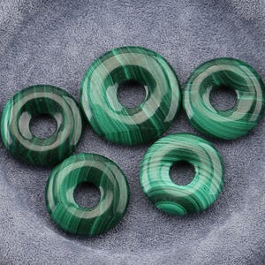 Puede incluir: Cinco piedras pulidas de malaquita en forma de rosquilla. Las piedras son de un verde vibrante con patrones arremolinados y un agujero central. La malaquita tiene un acabado brillante y está sobre un fondo gris.
