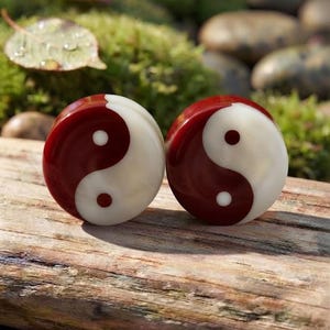 May include: A pair of circular ear plugs featuring a glossy yin and yang symbol design. Each plug displays a smooth, polished surface with a deep red and creamy white color scheme, showcasing the iconic swirling pattern with contrasting dots.