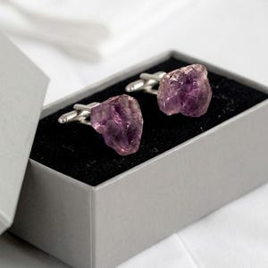 Puede incluir: Un par de gemelos con piedras de amatista moradas en bruto y sin pulir, unidas a herrajes de metal de tono plateado. Los gemelos se presentan dentro de una pequeña caja rectangular gris con un forro interior de color negro.