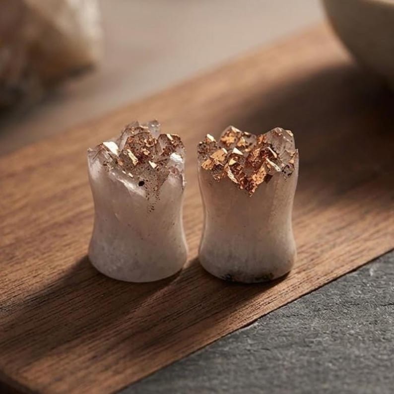 May include: A pair of cylindrical ear plugs made from off-white stone with a concave centre. The top edges of each plug feature a jagged, crystalline texture accented with metallic copper-coloured flakes embedded into the stone surface.