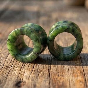 Op de afbeelding: Een paar cilindrische oorplugs gemaakt van gepolijste groene steen met natuurlijke donkere insluitingen en gevlekte patronen. Elk stuk heeft een gladde, cirkelvormige opening in het midden, die de gevarieerde textuur en diepe bosgroene tinten laat zien.