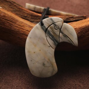 May include: A carved pendant in the shape of a bird's head, featuring a beak and eye details. The pendant is made of a stone with a marbled pattern of white, gray, and tan. It hangs from a black cord. A small, bone-colored piece is also visible.