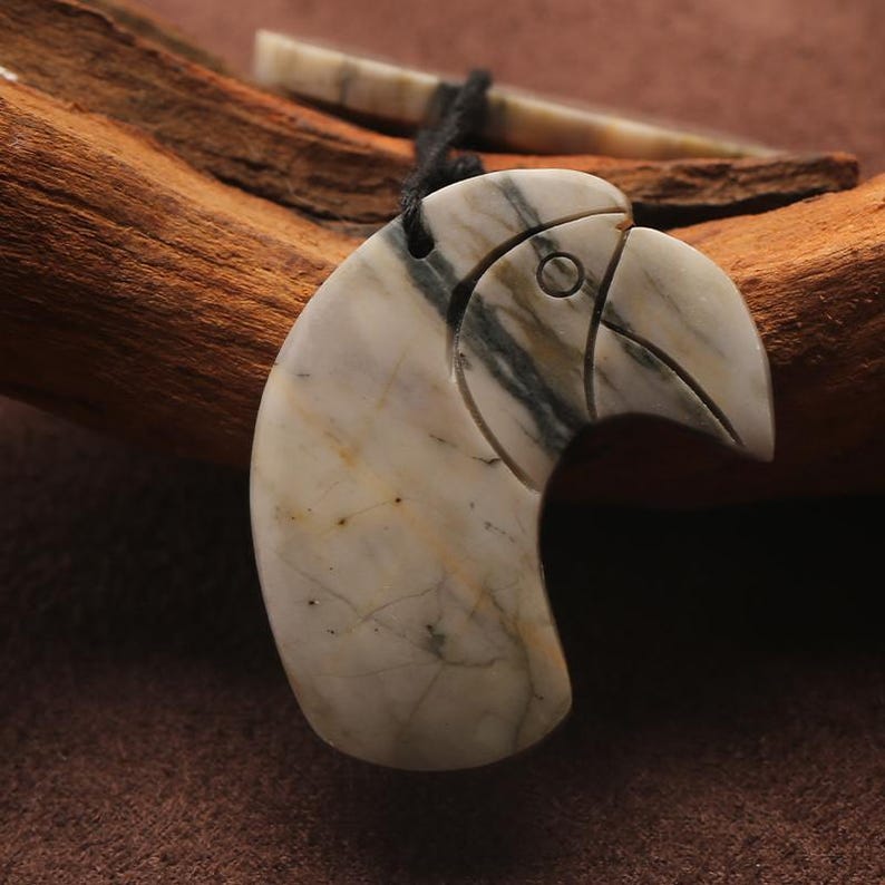 May include: A carved pendant in the shape of a bird's head, featuring a beak and eye details. The pendant is made of a stone with a marbled pattern of white, gray, and tan. It hangs from a black cord. A small, bone-colored piece is also visible.