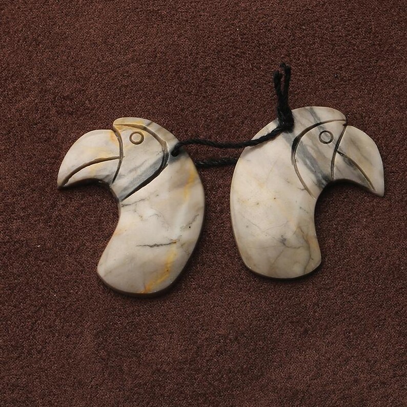 May include: Two carved stone pendants in the shape of bird heads, connected by a black string. The pendants are primarily off-white with gray and yellow veining. The background is a dark brown fabric.