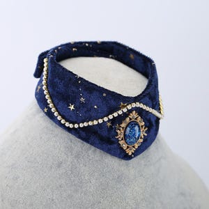 K&ouml;nnte beinhalten: Ein dunkelblauer Samtkragen mit goldenen Sternen und einem zentralen Schmuckstein. Der Kragen ist mit einer zarten Goldkette und einem gro&szlig;en, ovalen blauen Edelstein verziert, was ein elegantes Accessoire ergibt.