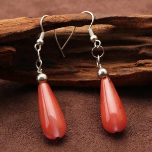 May include: A pair of drop earrings with coral-red, teardrop-shaped beads. The beads hang from silver-coloured hooks and small, round details. The earrings are set against a brown, textured backdrop.