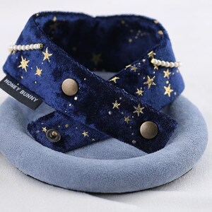 K&ouml;nnte beinhalten: Ein marineblauer Samt-Haustierhalsband mit goldenen Sternmustern und Perlenakzenten. Das Halsband hat einen Druckknopfverschluss und den Text "HONEY BUNNY" auf einem Etikett. Das Halsband liegt auf einer hellblauen kreisf&ouml;rmigen Basis.