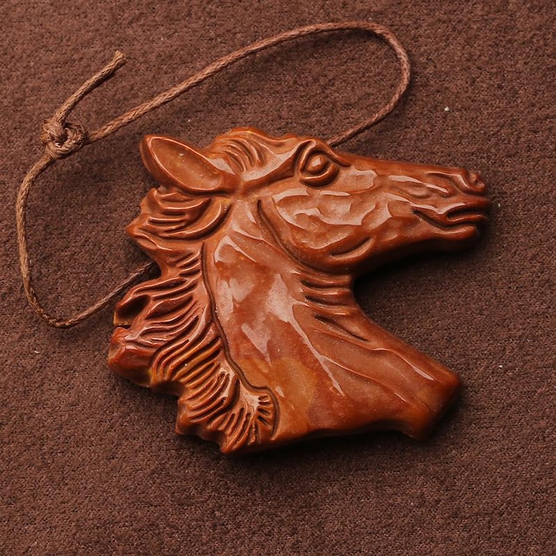 May include: A detailed, brown wooden carving of a horse's head, featuring intricate details of the mane and facial features. A brown string is attached for hanging. The carving is set against a brown background.