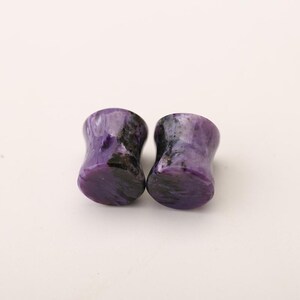 May include: A pair of cylindrical ear plugs crafted from polished purple stone with distinct black and dark violet swirling patterns throughout the material. The plugs feature a smooth, concave surface design with flared edges on both ends for secure placement.