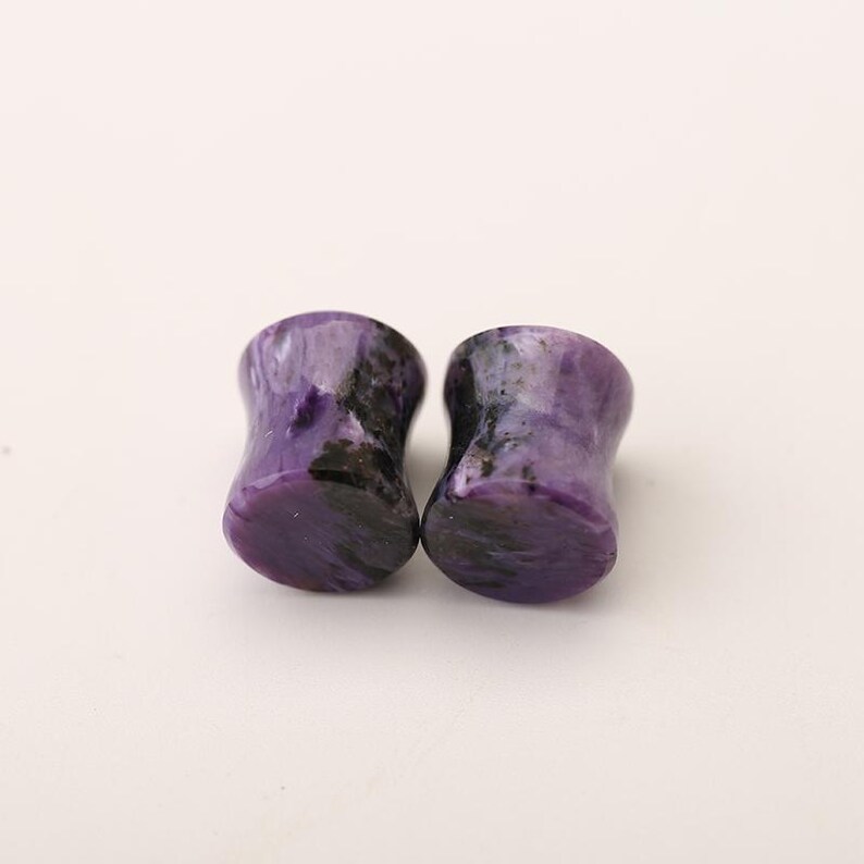 May include: A pair of cylindrical ear plugs crafted from polished purple stone with distinct black and dark violet swirling patterns throughout the material. The plugs feature a smooth, concave surface design with flared edges on both ends for secure placement.