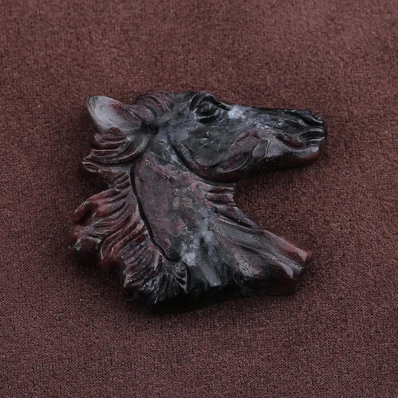 May include: A carved stone horse head sculpture in shades of black and burgundy. The sculpture is detailed, showing the horse's mane, eye, and ear. The background is a solid brown color.