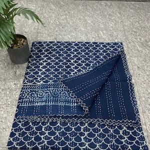 Op de afbeelding: Een gevouwen indigoblauwe quilt met een wit schubbenpatroon en decoratieve stiksels. De quilt heeft een contrasterende rand en is gemaakt van zachte stof. Een kleine potplant staat op de achtergrond.