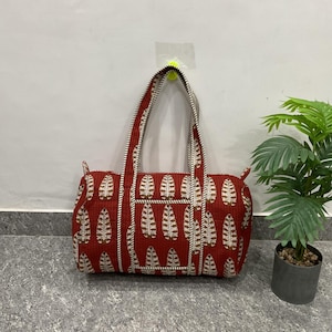 May include: A rectangular red and white patterned tote bag with two handles. The bag features a repeating leaf design in white against a red background. The handles are red with white and black stripes. A small potted plant is to the right.