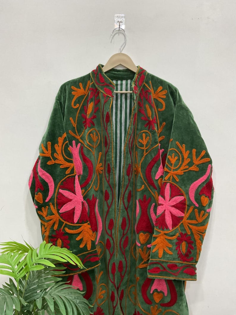K&ouml;nnte beinhalten: Ein gr&uuml;ner Samtmantel mit aufwendiger Blumenstickerei in Orange, Rot und Rosa. Der Mantel hat ein langes, offenes Design mit gestreiftem Futter und wird auf einem Holzb&uuml;gel pr&auml;sentiert.