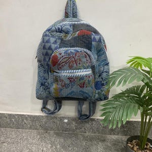 Puede incluir: Una mochila azul estampada con un pequeño bolsillo delantero y correas ajustables. La mochila está colgada en una pared. El estampado incluye diseños florales y geométricos en tonos azules, rojos y blancos.
