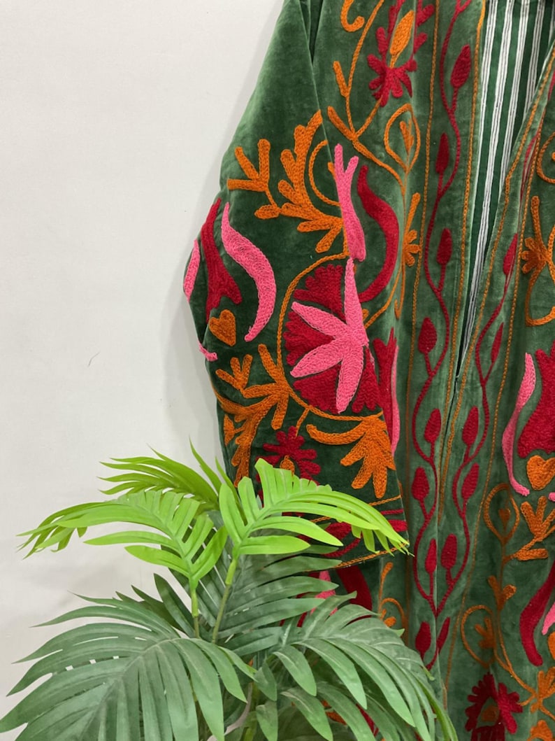 K&ouml;nnte beinhalten: Nahaufnahme eines gr&uuml;nen Samtgewands mit aufwendiger Stickerei. Die Stickerei zeigt ein Blumenmuster in Rot-, Orange- und Rosat&ouml;nen. Das Kleidungsst&uuml;ck hat ein gestreiftes Futter und eine lockere Passform.