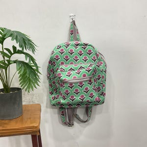 Puede incluir: Una mochila con base de tela verde y un patrón repetitivo de flores rosas con tallos de color marrón oscuro. La bolsa incluye un bolsillo frontal con cremallera, un asa superior y correas ajustables con un borde a rayas marrones y blancas.