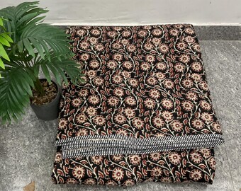 May include: Folded black fabric with a floral pattern in shades of red and cream. A section of the fabric is folded over, revealing a striped border in black and white. A potted plant is in the background.