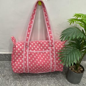 以下が含まれることがあります： ピンクと白のドット柄のトートバッグ。長方形の形で、2つのハンドルが付いています。ハンドルと縫い目は白です。柔らかい素材で作られています。