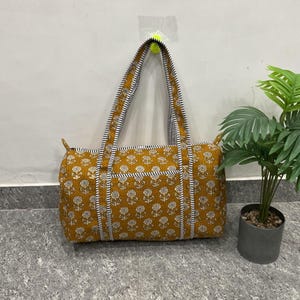 Puede incluir: Bolso tote de color amarillo mostaza con un estampado floral y ribete a rayas blancas y negras. El bolso tiene dos asas y un cierre de cremallera. El bolso mide aproximadamente 46 cm de ancho y 30 cm de alto.