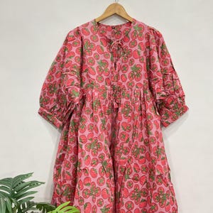 Könnte beinhalten: Ein knielanges Kleid mit rosafarbenem Hintergrund und einem sich wiederholenden Muster aus roten Tomaten mit grünen Blättern. Das Kleid hat Dreiviertelärmel mit gerafften Bündchen, einen Rundhalsausschnitt mit kleiner Bindung und eine lockere Passform.