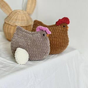 May include: Two plush, handmade chicken-shaped toys. One is brown with a red comb, the other is gray with a pink comb. A white, plush egg sits next to the gray chicken. A woven basket in the shape of a rabbit is in the background.