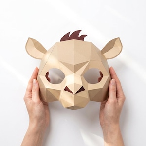May include: A low-poly paper mask in a light tan color, featuring a geometric animal design with pointed ears and a dark brown tuft of hair on top. The mask has two large circular eye openings and a triangular nose section, constructed from folded paper.