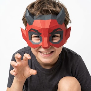 May include: A low-poly paper mask featuring a red face with black horns and pointed ears. The mask has angular geometric facets, cut-out eye openings, and a distinct devil-like design with sharp, defined lines across the forehead and nose.