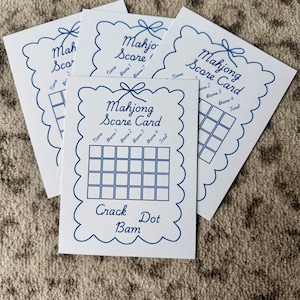 Könnte beinhalten: Ein Stapel weißer Mahjong-Scorecards mit blauen dekorativen Rändern und Text. Jede Karte hat ein Raster zum Ausfüllen, beschriftet mit "Name", "Spiel 1", "Spiel 2", "Spiel 3", "Spiel 4" und "Gesamt". Die Worte "Crack Dot Bam" stehen unten.