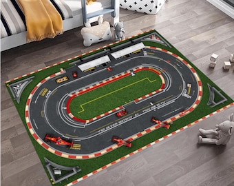 May include: A rectangular children's play rug featuring a race track design. The rug has a green interior with a gray track, red and white borders, and toy race cars. The rug is placed on a wooden floor.