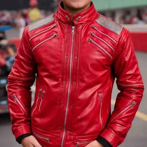 Può includere: Giacca in pelle rosso vivo con multiple cerniere argentate e colletto alto. La giacca presenta dettagli argentati sulle spalle ed è abbinata a jeans blu. Un capo di moda audace.