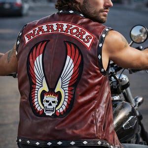 May include: A burgundy leather vest with the word "WARRIORS" across the back. The vest features a large patch with a skull wearing a crown, wings, and red, white, and yellow accents. The vest is worn over a black shirt and jeans.