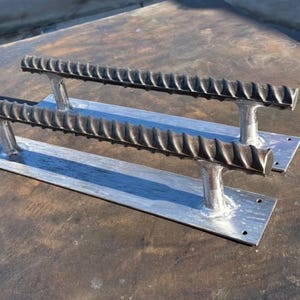 May include: Two metal door handles with a rebar design. Each handle has a rectangular base with two vertical supports holding a textured, cylindrical bar. The handles are silver and sit on a dark, textured surface.