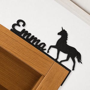 Może przedstawiać: Czarna, brokatowa dekoracja narożna w kształcie litery L, przedstawiająca imię Emma napisaną kursywą oraz sylwetkę stojącego jednorożca. Element jest przeznaczony do umieszczenia na górnym rogu drewnianej ościeżnicy drzwiowej.