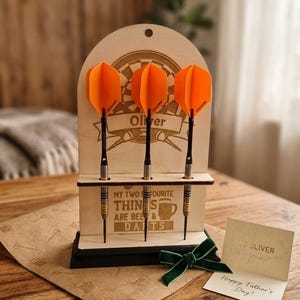 Könnte beinhalten: Ein hölzerner Dartständer mit drei Darts, jeweils mit leuchtend orangefarbenen Flights. Der Ständer trägt den Namen "Oliver" und den Text "MY TWO FAVOURITE THINGS ARE BEER & DARTS". Eine "Happy Father's Day"-Karte ist ebenfalls vorhanden.