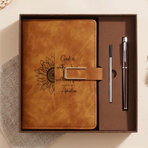May include: A tan leather-like journal with a sunflower illustration and the text God is within her she will not fall Psalm 46:5 Amelia, featuring a gold-tone metal clasp, presented in a gift box with a matching metal pen and a refill ink cartridge.