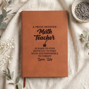 May include: A tan leather-bound notebook featuring the black engraved text: A TRULY DEVOTED Math Teacher IS HARD TO FIND DIFFICULT TO PART WITH AND IMPOSSIBLE TO FORGET Love, Lily. The notebook includes a small inkwell illustration below the word Math.