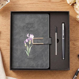 Op de afbeelding: Een donkergrijs notitieboek met een goudkleurige metalen sluiting, voorzien van een paarse irisillustratie en de tekst "THE NEXT Chapter BEGINS" en "CHOLE". Het notitieboek zit in een zwarte geschenkdoos met een bijpassende grijze pen en vulling.