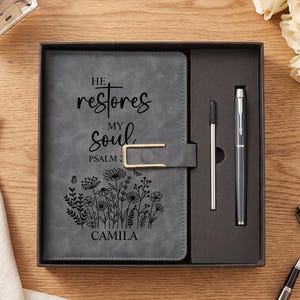Op de afbeelding: Een grijs notitieboek met een goudkleurige metalen sluiting, voorzien van de tekst "HE restores MY soul PSALM 23" en "CAMILA" boven een zwarte bloemenillustratie. Het notitieboek zit in een donkere doos met een bijpassende grijze pen en vulling.