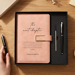Könnte beinhalten: Ein pfirsichfarbenes Notizbuch mit einem goldfarbenen Metallverschluss, auf dessen Einband der Text "The next chapter" und "AMELIA SMITH" gedruckt ist. Das Notizbuch befindet sich in einer schwarzen Geschenkbox mit einem schwarzen Stift und einer silbernen Mine.