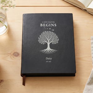 May include: A black hardcover journal featuring a white graphic of a tree with exposed roots. The text reads A NEW CHAPTER BEGINS, Daisy, and EST 2026. A thin brown ribbon bookmark extends from the bottom edge of the rectangular book cover.