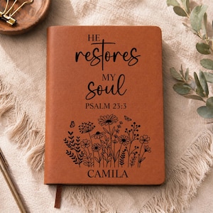 Op de afbeelding: Een beige journal in lederlook met zwarte gravure met de tekst HE restores MY soul PSALM 23:3 en CAMILA. Onder de tekst staat een zwarte lijntekening van diverse wilde bloemen en twee kleine vlinders.