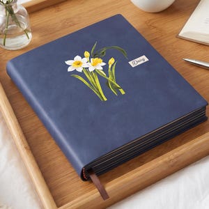 May include: A navy blue hardcover journal featuring a botanical illustration of white and yellow daffodils on the front cover. A small rectangular label with the word Daisy printed in black text is positioned to the right of the floral graphic.