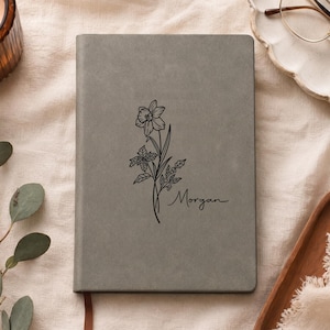 Könnte beinhalten: Ein graues Notizbuch mit einer schwarzen Strichzeichnung einer Narzissenblüte und dem Namen Morgan in Schreibschrift. Der Einband hat eine glatte, matte Textur und eine rechteckige Form mit abgerundeten Ecken auf einem hellen Stoff.