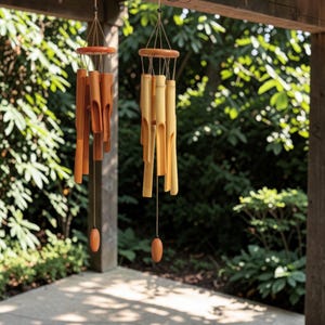 May include: Two bamboo wind chimes hanging outdoors. One is a light brown color, and the other is a darker brown. Each chime has a wooden top and a wooden oval-shaped weight at the bottom. The background is a lush green garden.