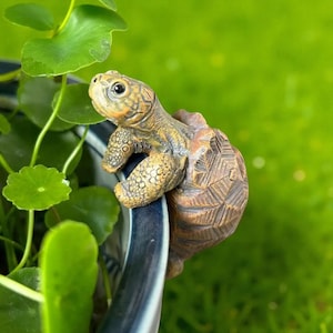 May include: A detailed, brown and tan turtle figurine climbing over the edge of a blue and white ceramic pot. The turtle has textured skin and is surrounded by green foliage and a blurred green background.